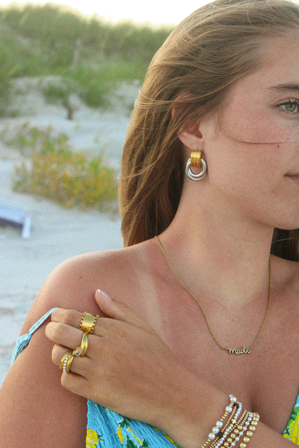 Woman on a beach wearing gold jewelry including earrings, rings, and bracelets.