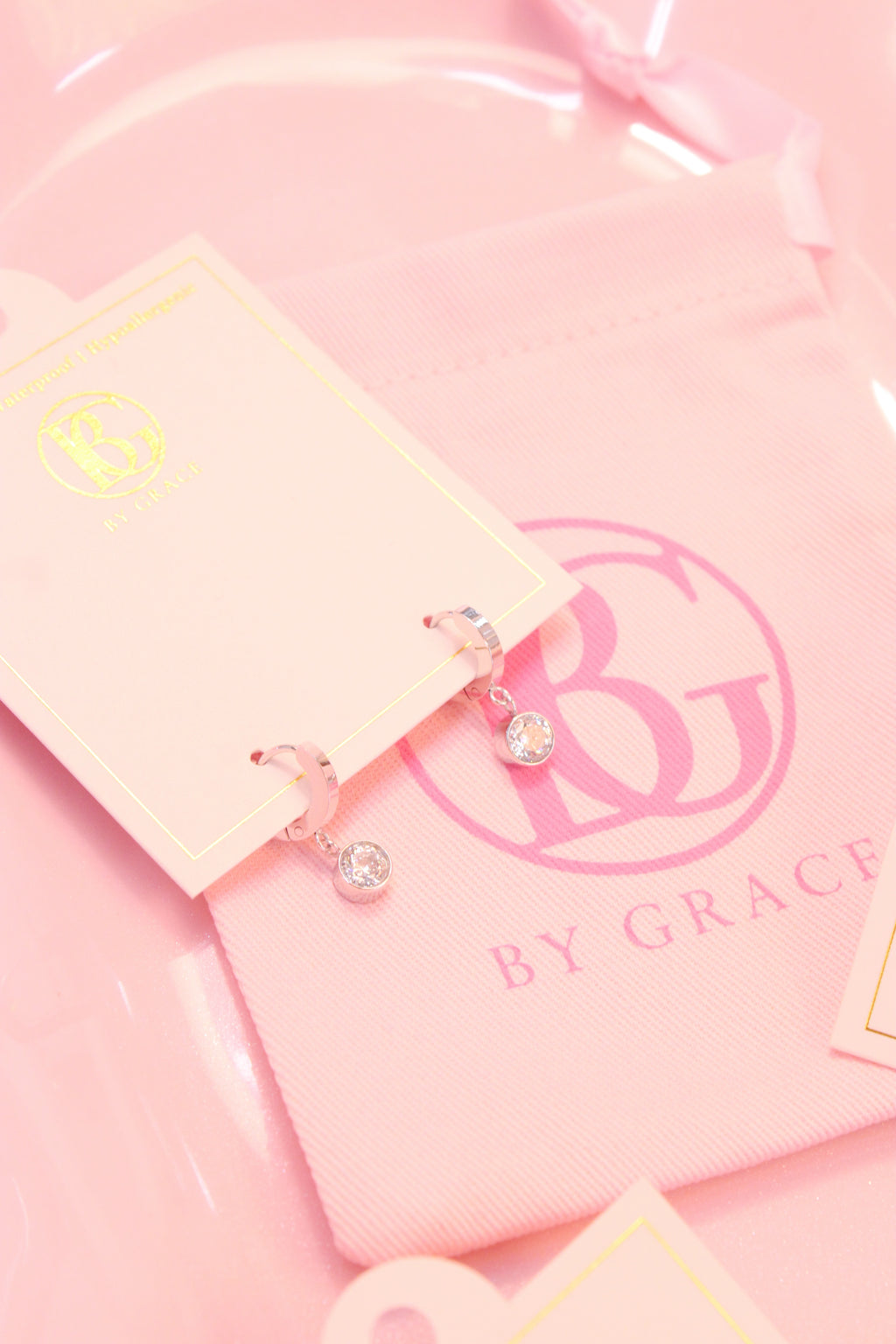Pink jewelry card with silver hoop earrings with small drop diamond  and 'BY GRACE' branding on a pink background