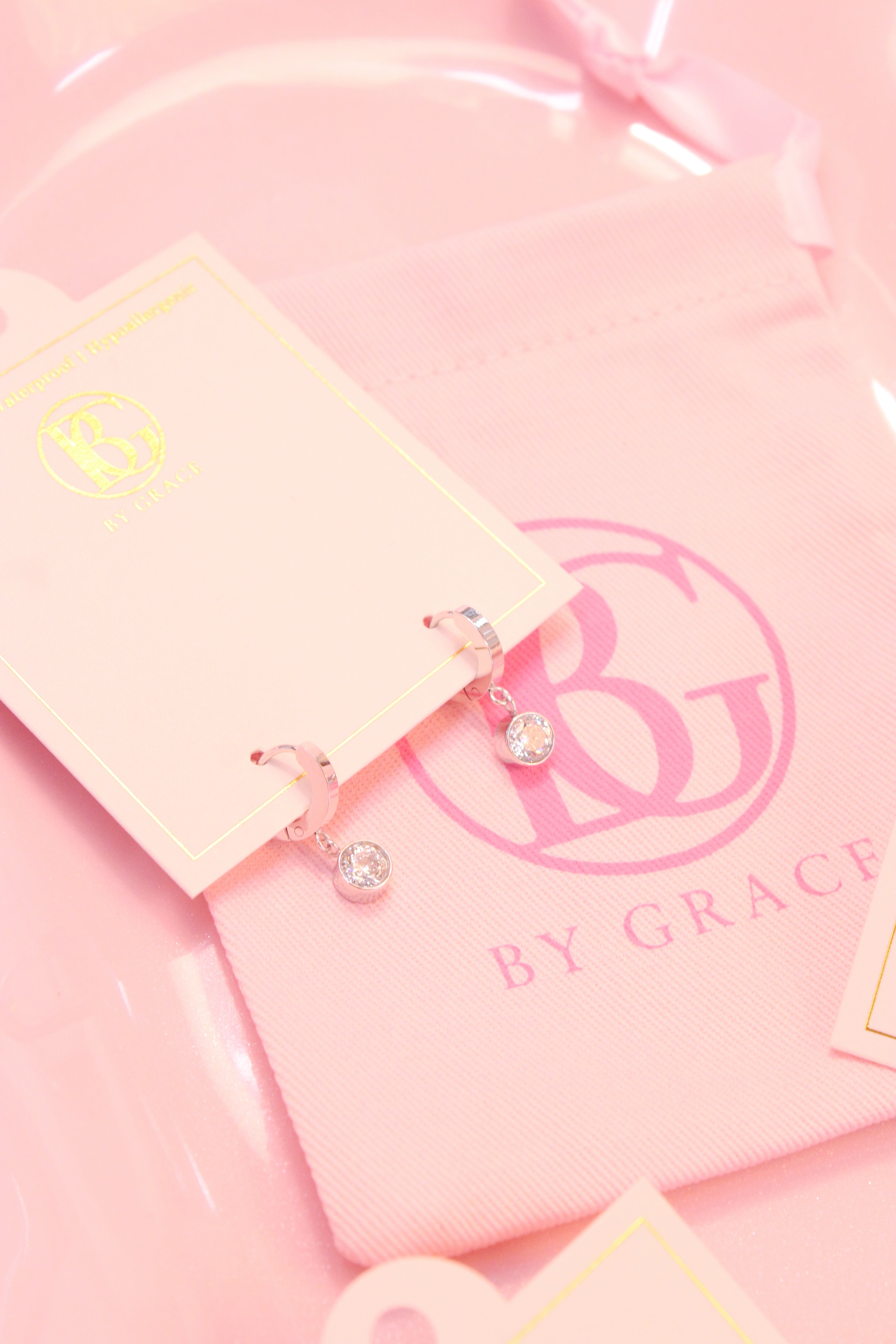 Pink jewelry card with silver hoop earrings with small drop diamond  and 'BY GRACE' branding on a pink background