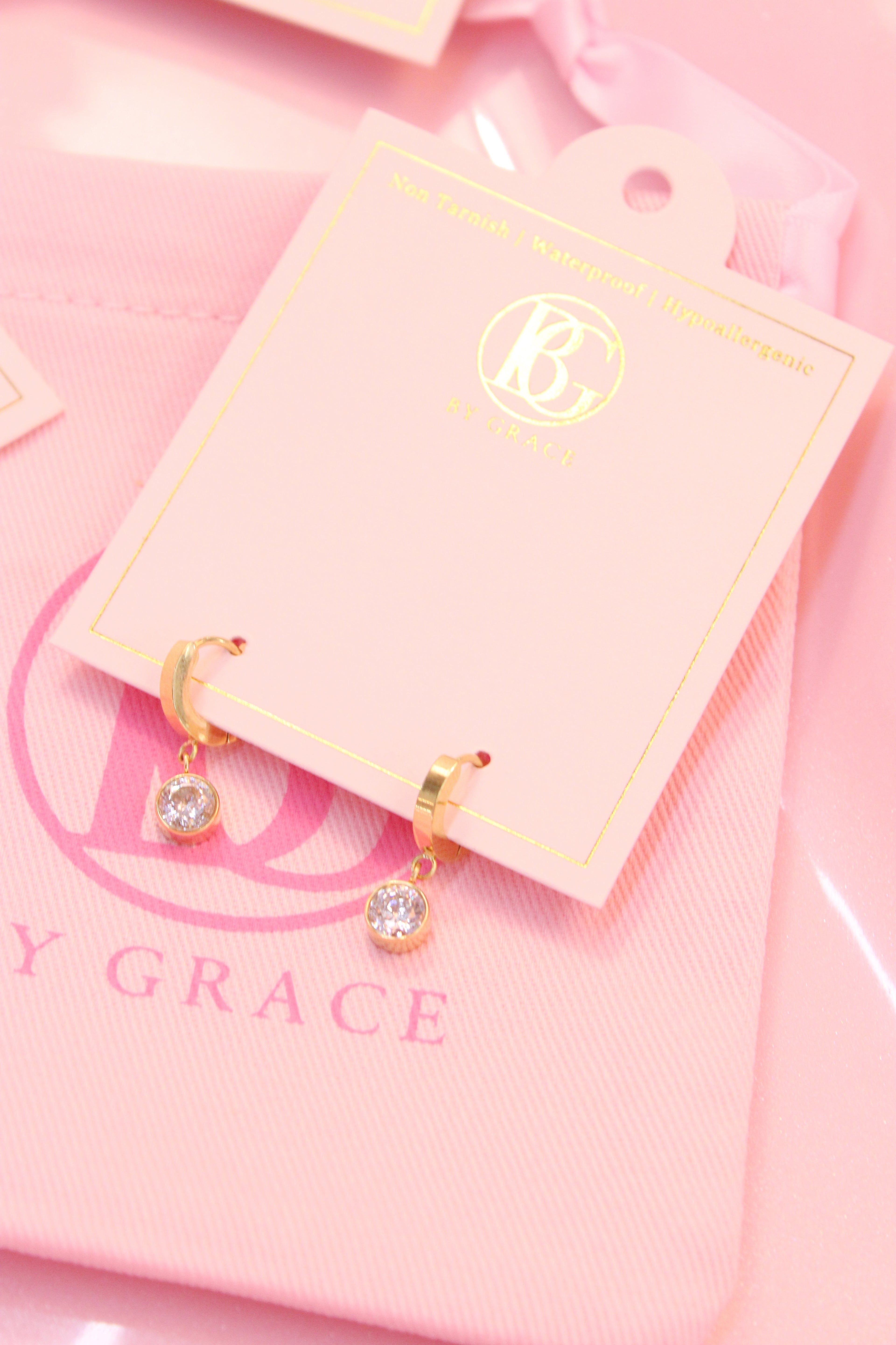 Gold hoop earrings with small drop diamond on a pink card with 'By Grace' branding.