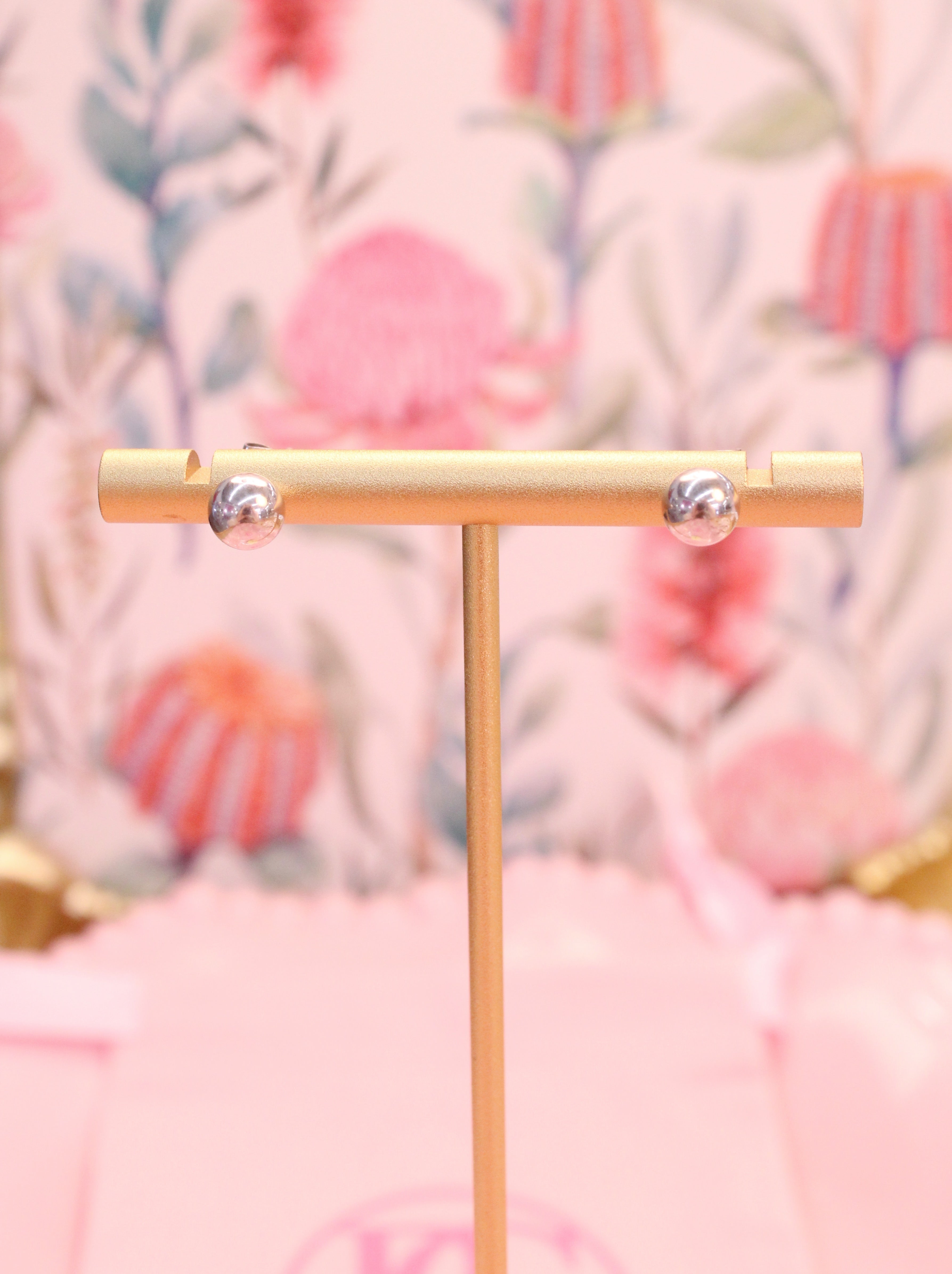 Gold T-shaped stand with silver ball earrings against a textured pink and orange background 